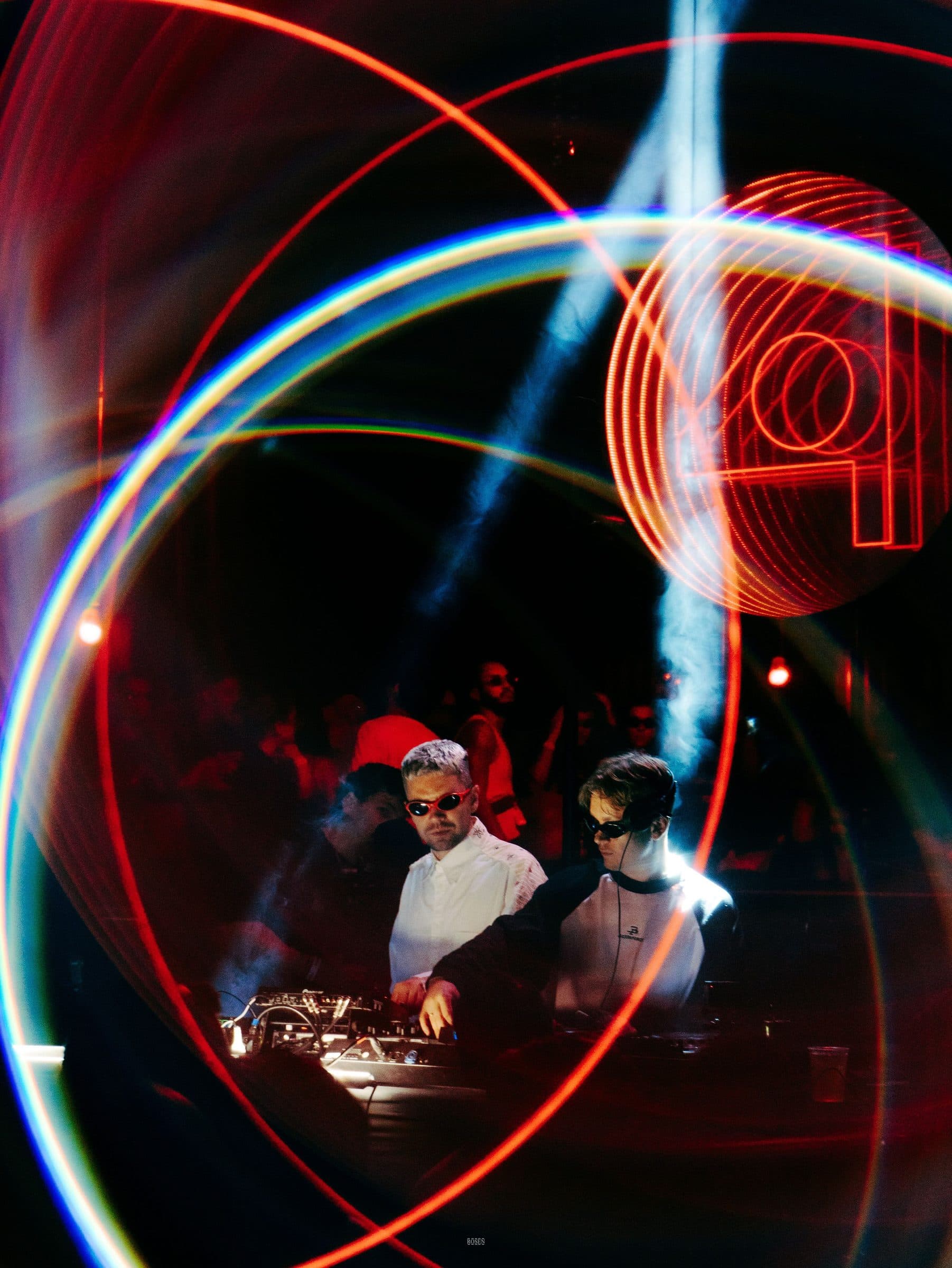 Two DJs at the decks inside rings of red and blue light, a crowd behind them